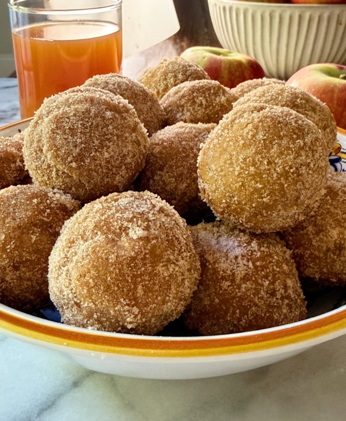 Bowl filled with baked gluten-free apple cider donut holes coated in cinnamon sugar, with fresh apples and a glass of apple cider in the background.